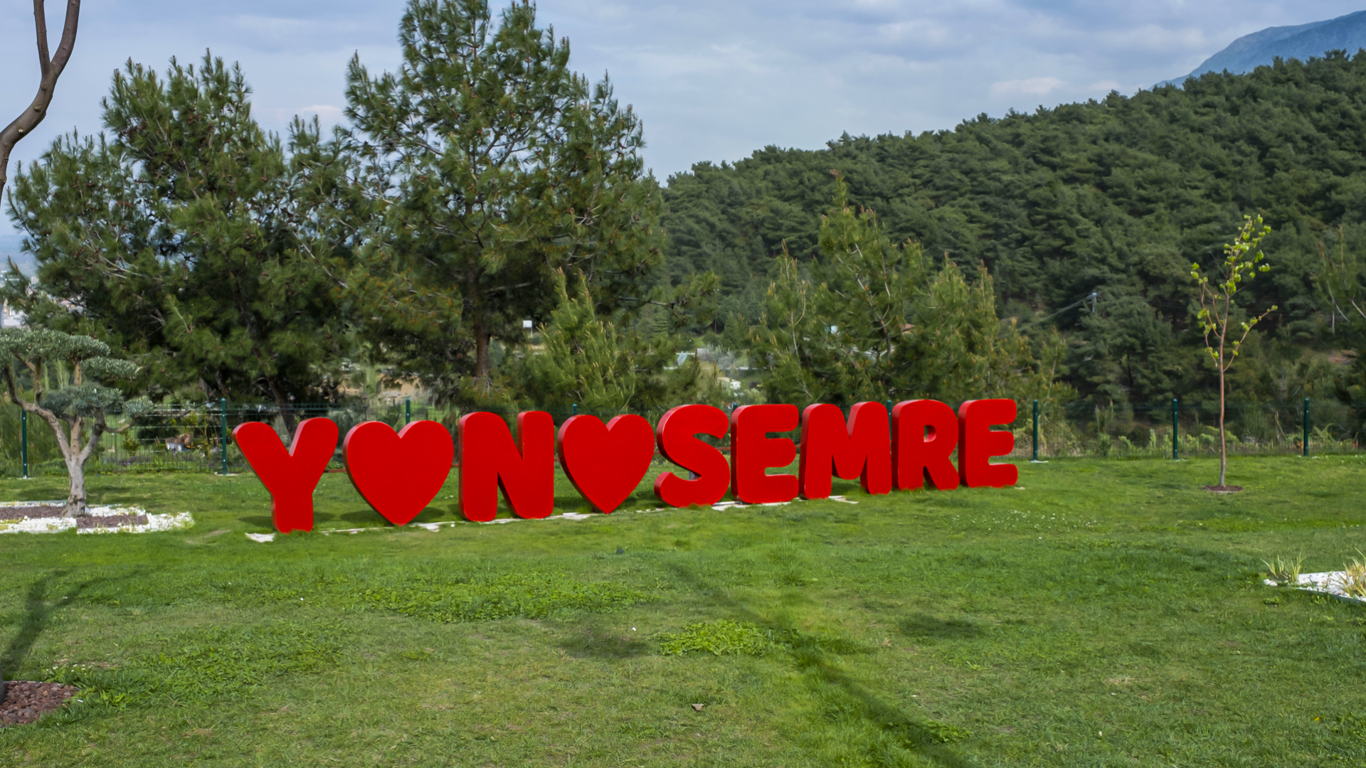 Yemyeşil ormanlık bir tepenin önündeki geniş çimenlik alanda yer alan, kalp figürleri içeren kırmızı renkli devasa "YUNUSEMRE" yazısını göstermektedir.