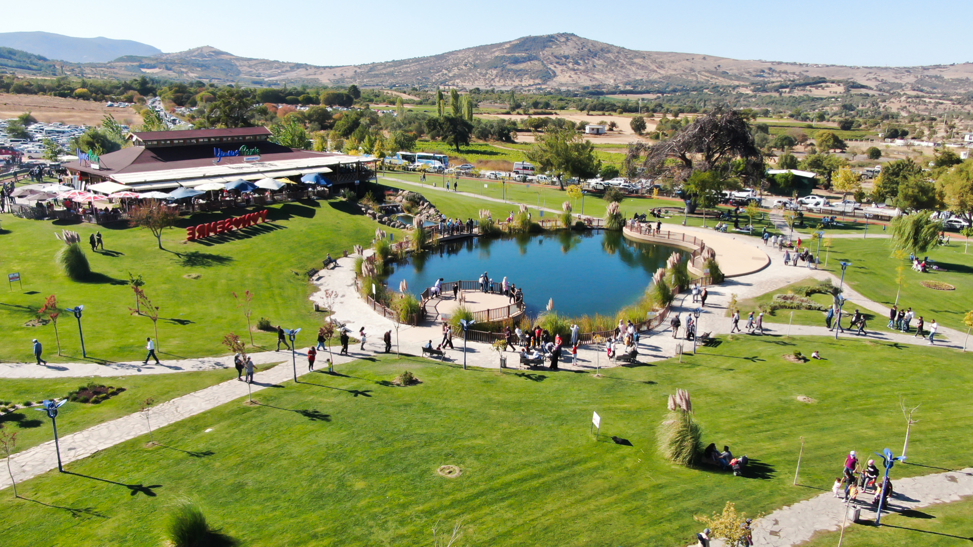 Ortasında masmavi bir gölet bulunan, etrafı yürüyüş yolları ve geniş çim alanlarla çevrili, arka planında dağların yer aldığı güneşli bir park manzarasını yukarıdan göstermektedir.