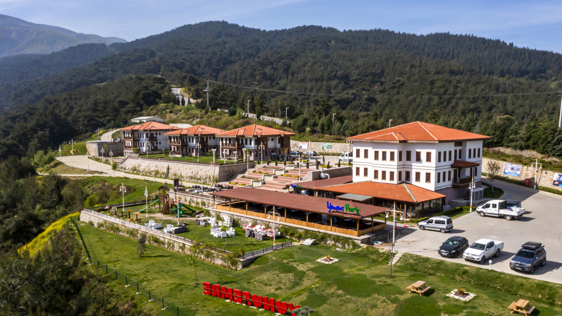 Ormanlık bir dağın yamacında, geniş yeşil alanlar içine inşa edilmiş kırmızı kiremit çatılı büyük bir tesis kompleksini göstermektedir.